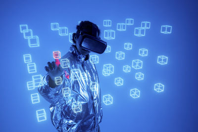 Woman wearing virtual reality simulator touching hologram cube on screen against blue background