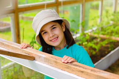 Pretty little girl in a white panama, looks out of the window of a wooden vegetable greenhouse