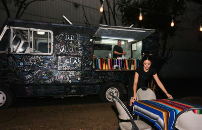 Ethnic lady covering table on street while male colleague working at counter of food truck at nighttime