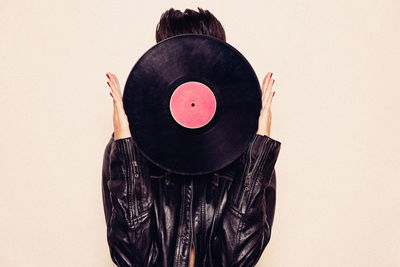 Faceless woman in leather jacket hiding face behind vinyl record in studio on pink background