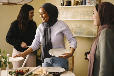 Smiling young woman arranging plates at table while standing by friends in kitchen