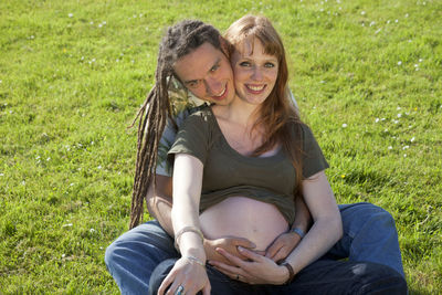 Portrait of happy man embracing pregnant girlfriend sitting on grassy field