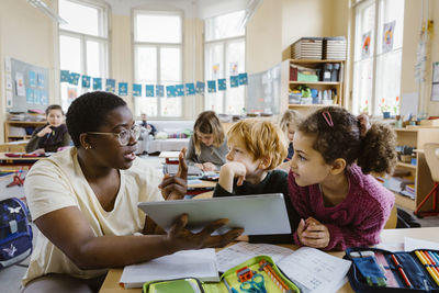 Teacher with tablet pc explaining schoolboy and schoolgirl at desk while gesturing in classroom