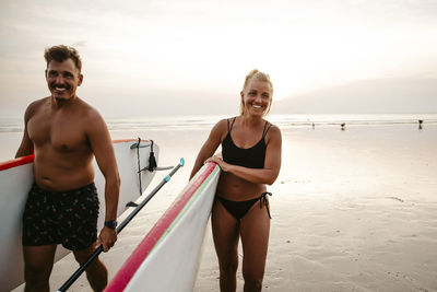 Happy couple with paddleboards walking at beach during sunset