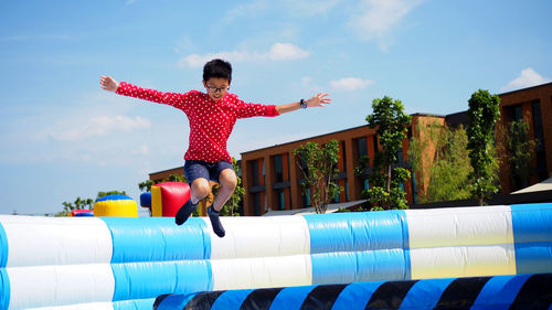 Full length of boy with arms outstretched jumping on bouncy castle against sky