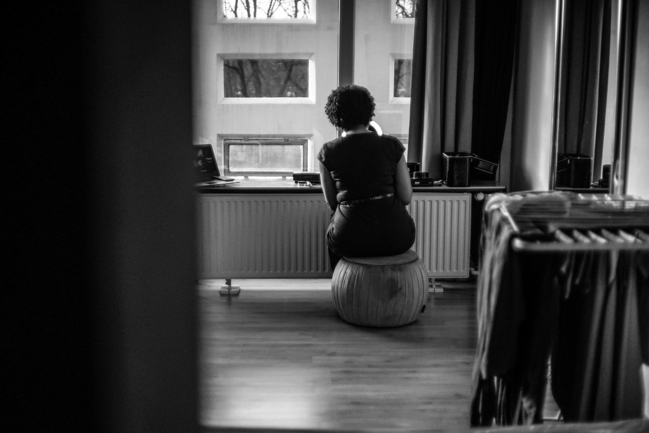 one person, rear view, indoors, sitting, full length, real people, window, lifestyles, home interior, seat, domestic room, adult, women, solitude, leisure activity, standing, men, young adult, waiting, hairstyle, contemplation