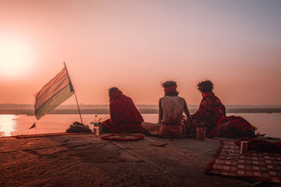 Rear view of people sitting by sea against sky during sunset