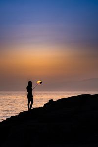 Silhouette woman taking selfie using smart phone and monopod at beach against sky during sunset