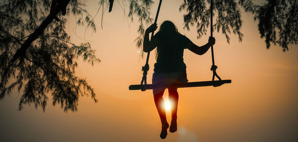 Rear view of girl sitting on swing at beach against sky during sunset