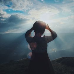Rear view of woman shielding eyes while standing on cliff against cloudy sky