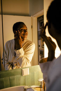 Smiling young woman applying moisturizer on face while looking in mirror at home