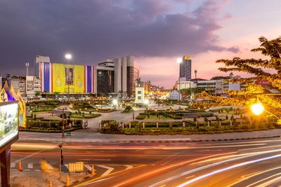 Light trails on road by buildings against sky during sunset