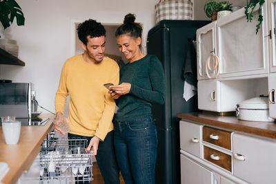 Woman smiling while showing smart phone to boyfriend while standing in kitchen