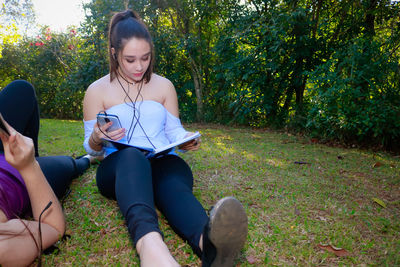 Young woman with book by friend listening to music on grassy field