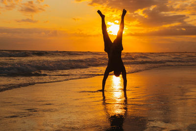 Rear view of woman with arms outstretched standing at beach against sky during sunset