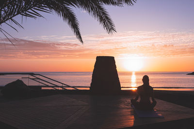 Young woman's silhouette meditating at sunrise in front of the beach.