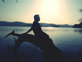 Silhouette man relaxing on log at lakeshore against sky during sunset