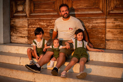 Dad sitting with small baby and two elder children on the stairs against wooden door