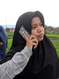 Cropped hand of boy holding mobile phone for sister talking while standing against clear sky