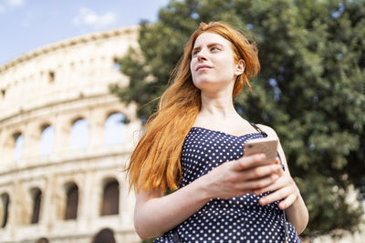 Dreamy ginger tourist using smartphone near famous attraction