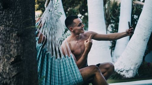 Shirtless young man taking selfie with smart phone while sitting on hammock