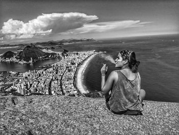 Young woman smoking cigarette while sitting over sea against sky