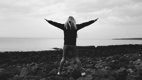 Rear view of woman with arms outstretched standing at beach against sky