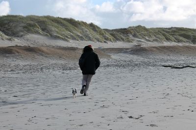 Rear view of mid adult man playing with dog while walking at beach against sky