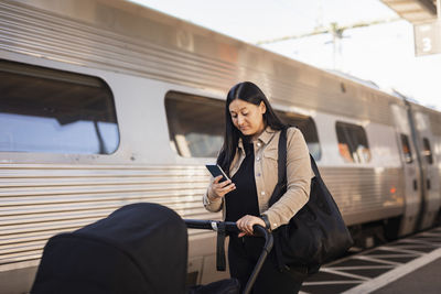 Mid adult woman at train station pushing pram and using cell phone