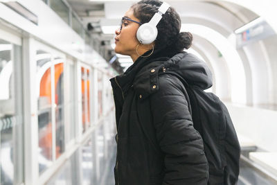 Side view of relaxed colombian female passenger in outwear and backpack listening to songs from playlist in wireless headphones while waiting for train on underground station
