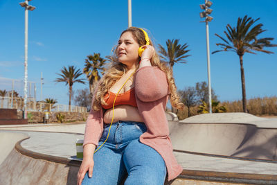 Young woman listening music sitting against sky