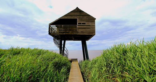 Boardwalk leading towards stilt house by sea against sky