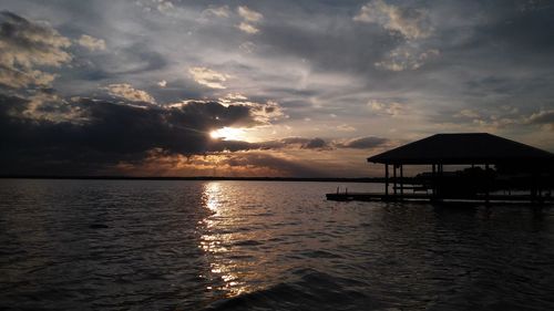 Silhouette gazebo by river against cloudy sky during sunset