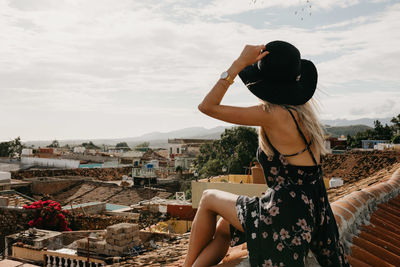 Side view of young woman looking at cityscape while sitting on rooftop against sky during sunny day