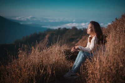 Side view of woman sitting on high field against sky