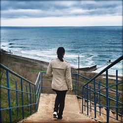 Full length rear view of woman walking on steps leading towards sea against sky