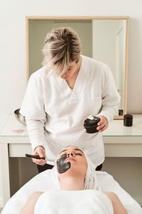 Professional cosmetician applying black anti aging mask on face of female client during skin care procedure in modern beauty salon