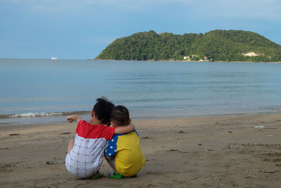 Rear view of brothers sitting at beach against sky