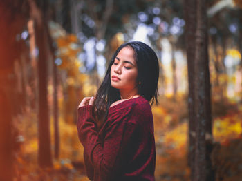 Young woman looking away while standing against trees during autumn
