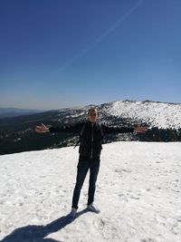 Portrait of young man with arms outstretched standing on snowcapped mountain against clear sky during sunny day
