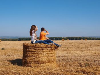 Side view of couple sitting on hay bale against clear sky