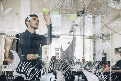 Young creative businessman sticking adhesive note on patterned glass in office