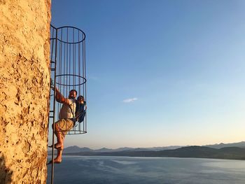 Father climbing ladder on wall while carrying son against sky