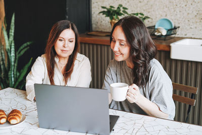 Adult smiling middle aged women sisters friends having breakfast and using laptop in kitchen 