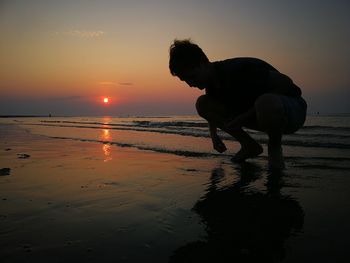 Boy crouching at beach against sky during sunset