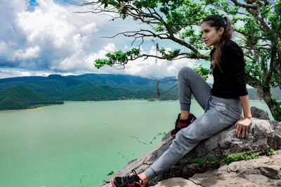 Young woman sitting by tree against sky