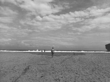 Rear view of young man walking at beach against cloudy sky