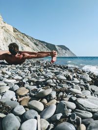 Shirtless man lying on rocks at beach against clear sky