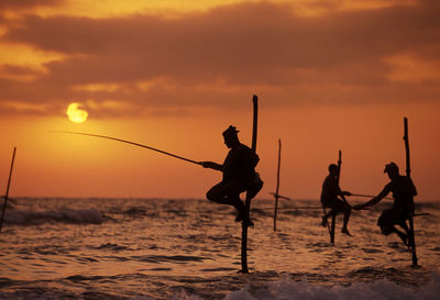 Silhouette fishermen stilt fishing in sea during sunset