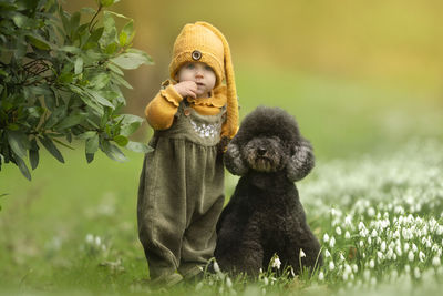 Little girl in cute dress is standing outdoors in nature with snowdrops with a black poodle